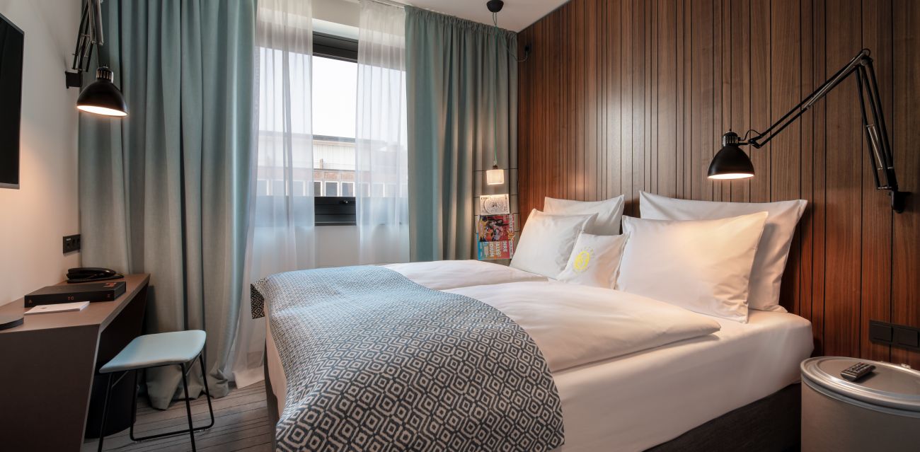 View into SMALL room with bed and desk in the foreground, behind it a window